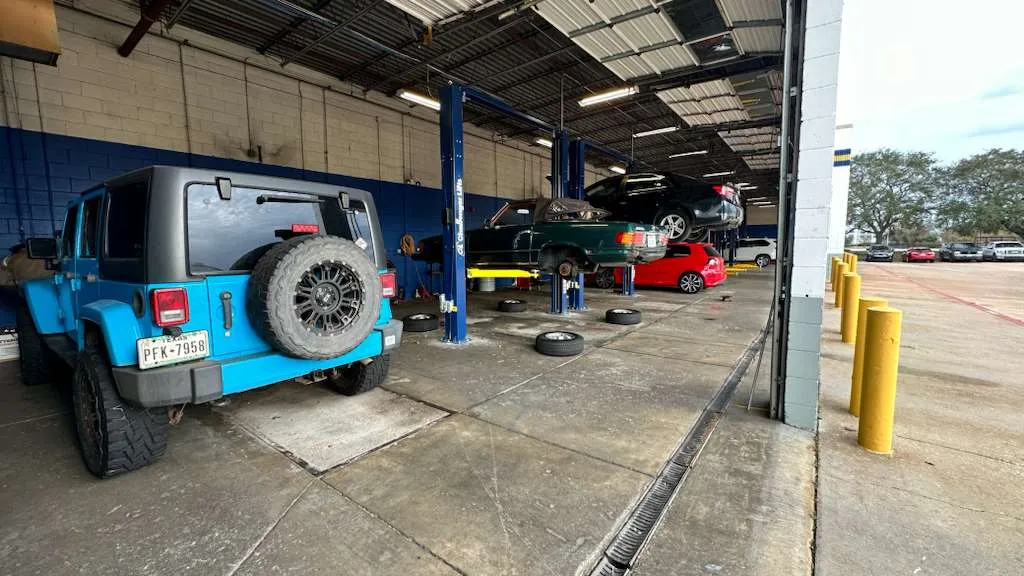 Classic green Mercedes-Benz R107 on a hydraulic lift in a professional auto repair shop with tires removed for service.