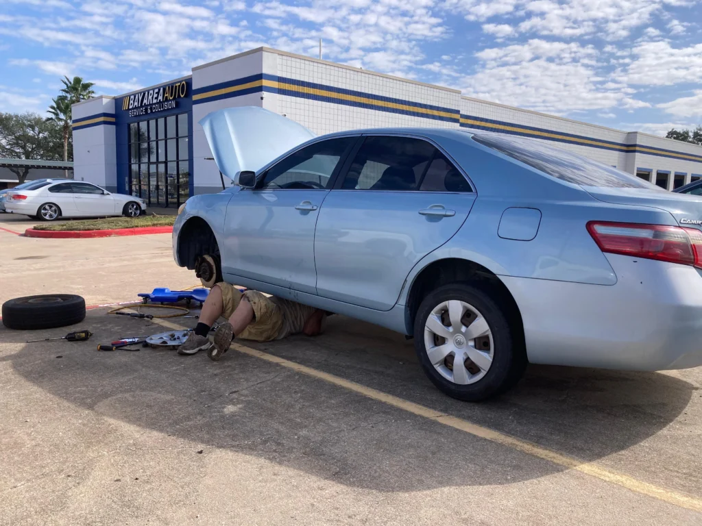 Professional mechanic performing auto repair under a blue sedan at Bay Area Auto service and collision center.