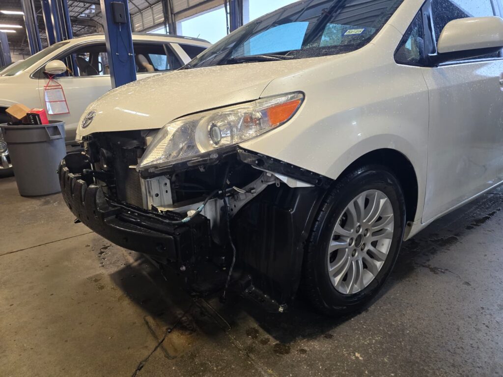 White sedan with significant front-end collision damage showing crushed bumper, bent frame, and exposed headlight assembly during professional inspection in collision repair shop