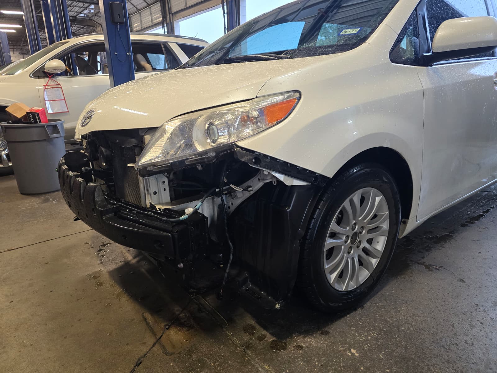 White sedan with significant front-end collision damage showing crushed bumper, bent frame, and exposed headlight assembly during professional inspection in collision repair shop
